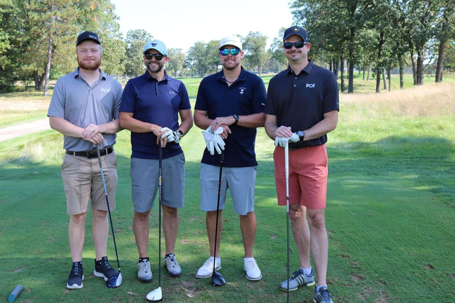 Four people stand in a line on a golf course, wearing polo shirts and hats, holding golf clubs and smiling at the camera.