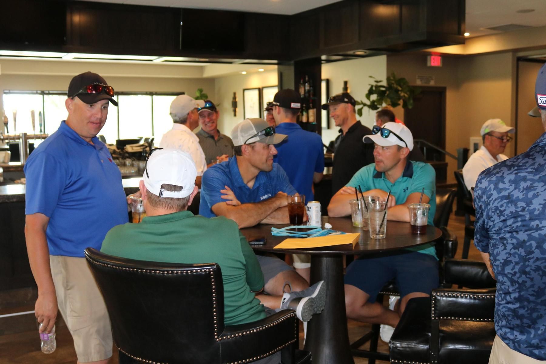 A group of people wearing casual clothing and hats gathered around a circular table in a clubhouse setting.