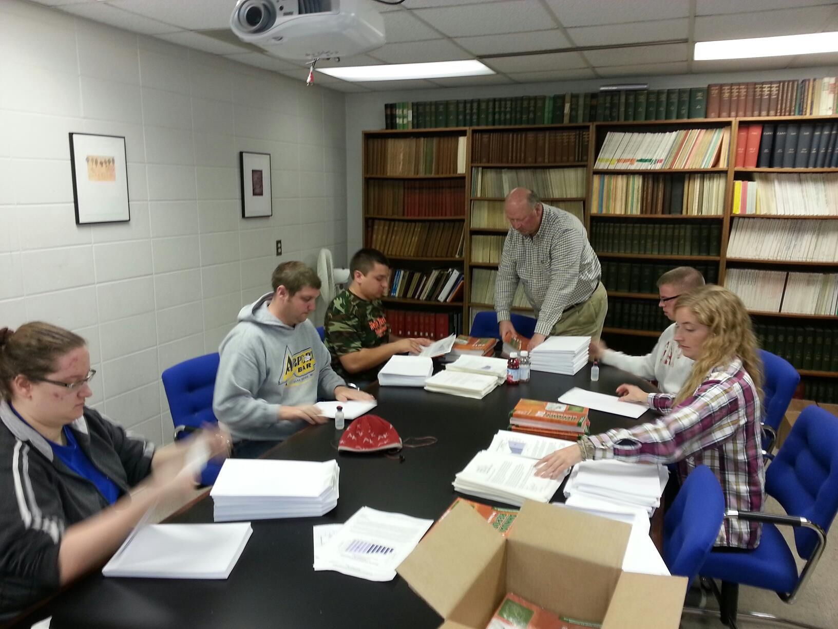 Five people sit around a table in a library setting, working together on stacks of documents.