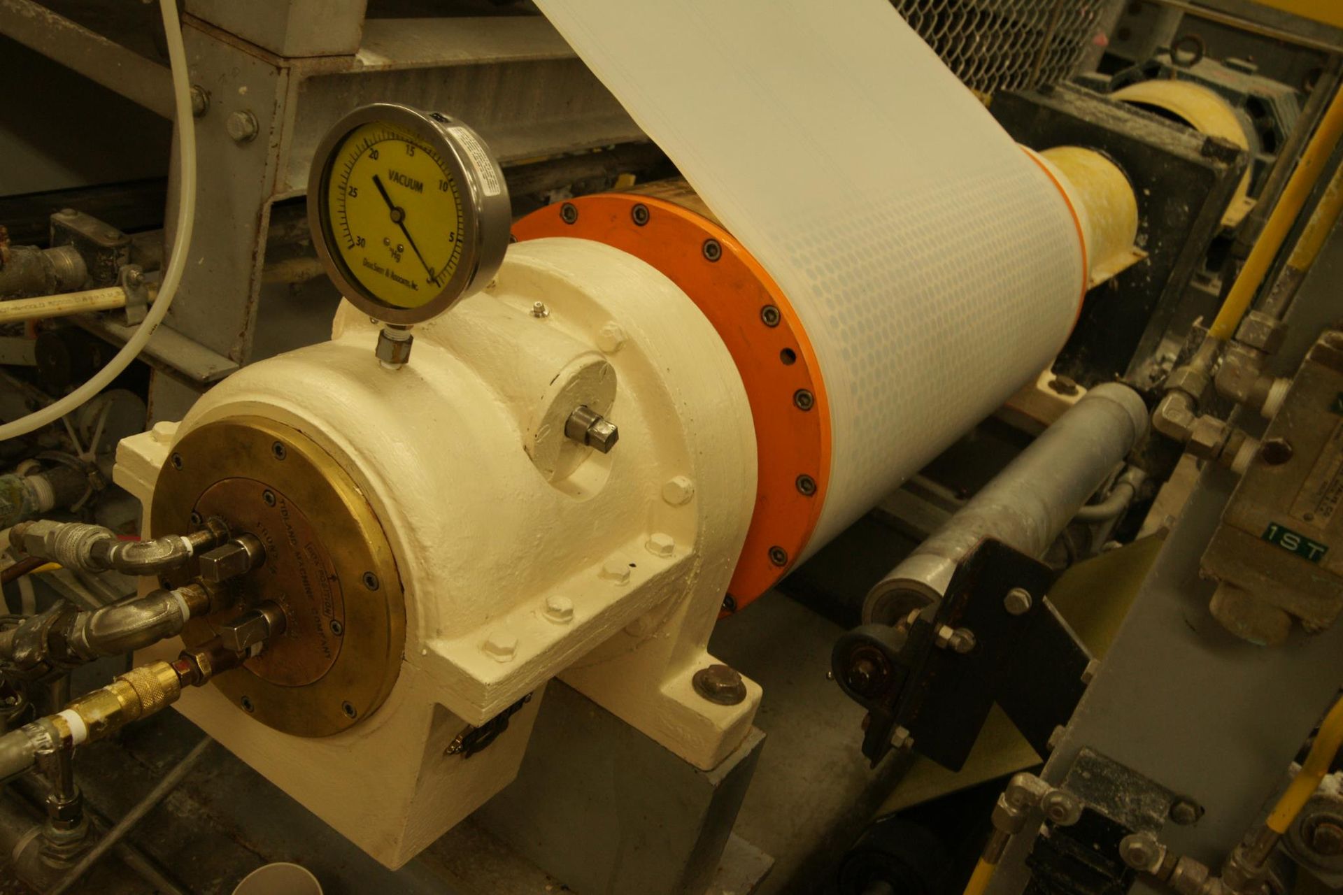 A white industrial machine roller with an orange flange and a pressure gauge, processing a white sheet of material.