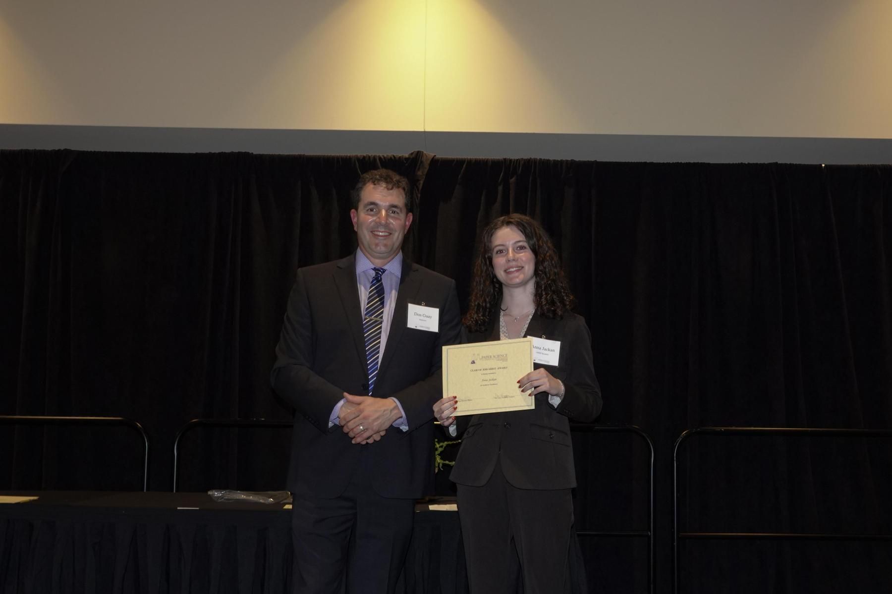 A person in a suit stands next to another person holding a certificate at a podium in front of a black curtain.