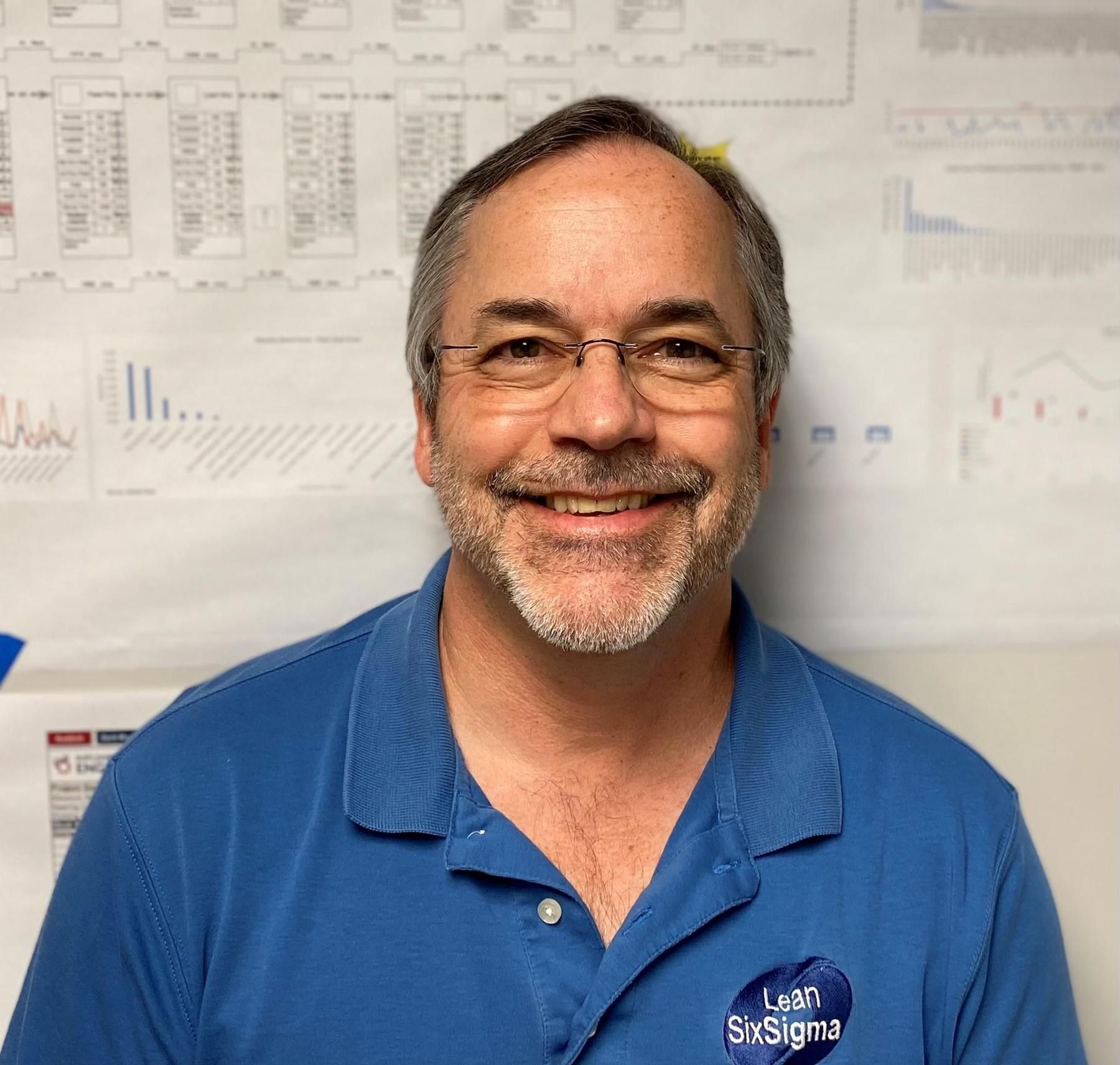 A person with a beard and glasses wearing a blue polo shirt, smiling in front of a white wall with charts and diagrams.