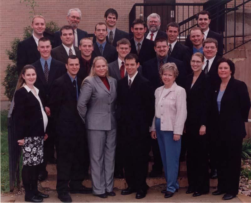 A group of twenty people in professional attire posing for a formal photo outside a brick building with metal stairs.