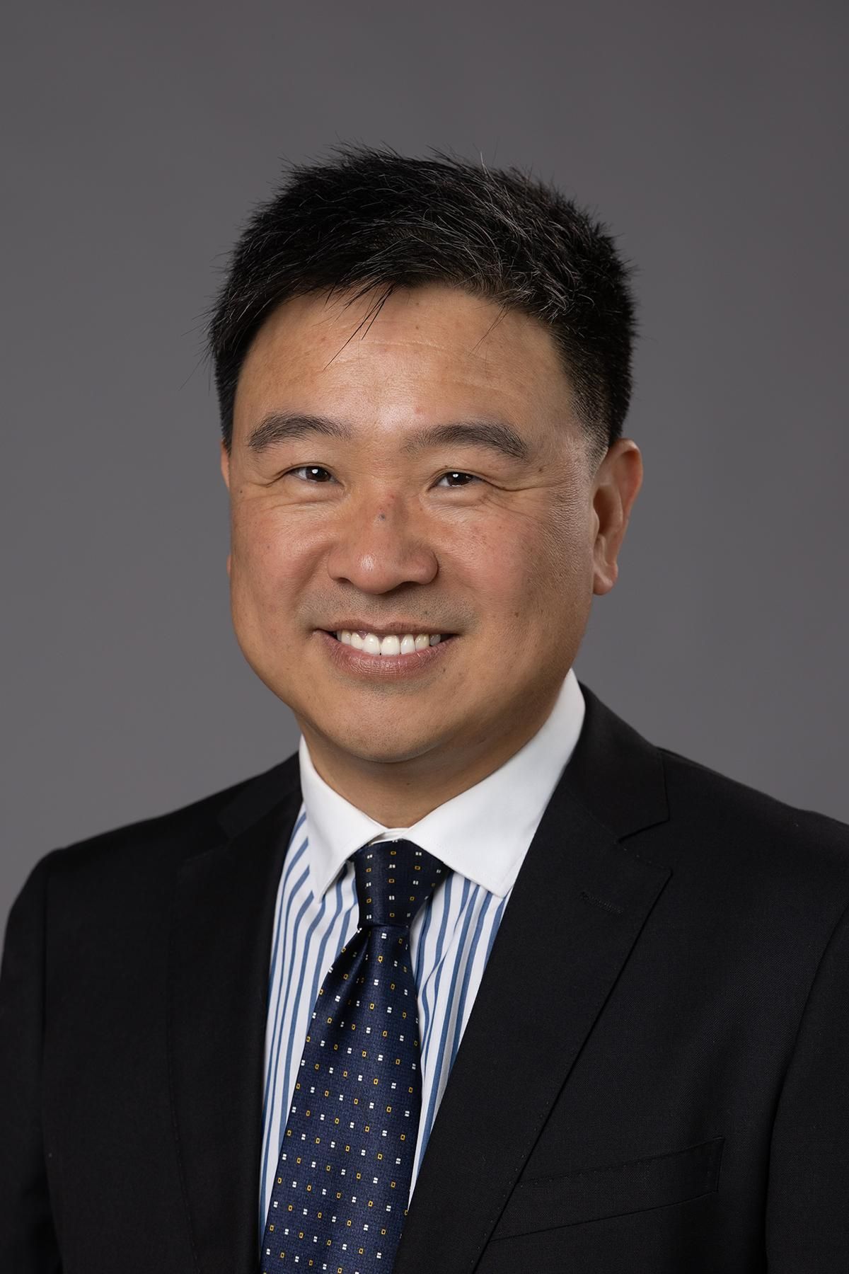 A professional headshot of a smiling person in a dark suit, light-blue striped dress shirt, and dark blue polka-dot tie.