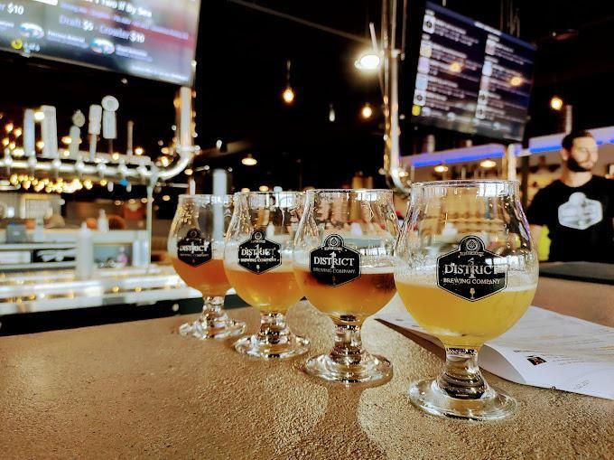 A flight of four glasses of golden beer arranged in a row on a bar counter inside a brewery.