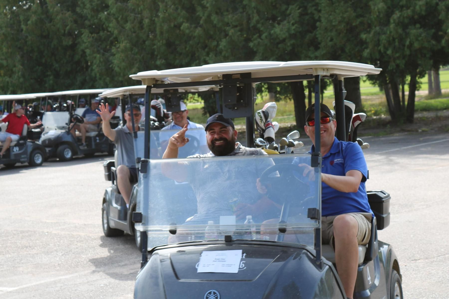 People in golf carts on a parking lot at a golf course, with some waving and smiling.