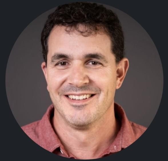 A portrait of a smiling person with curly dark hair wearing a collared reddish shirt against a plain dark gray background.