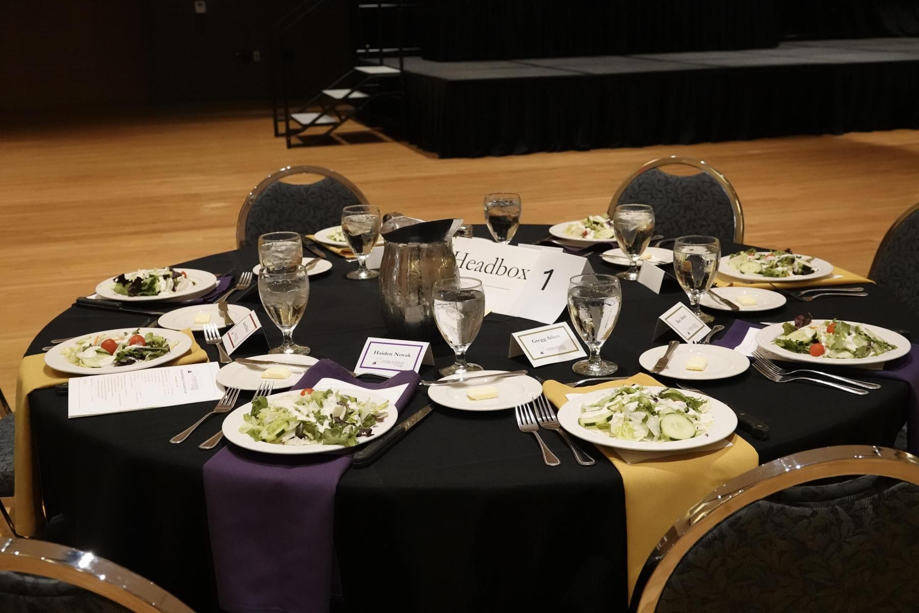A round banquet table with black tablecloth, salads, glasses, and purple and gold accents in an event space.