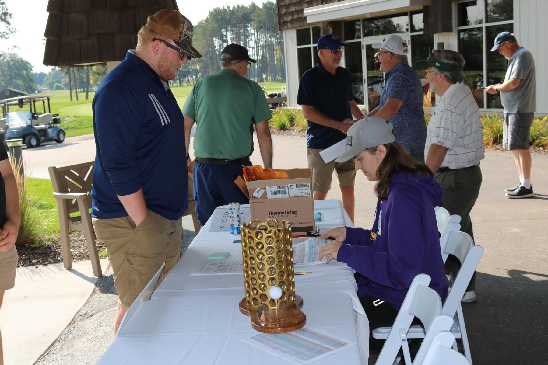People gather at an outdoor registration table with a golden trophy and a cardboard box in a parking area near a golf course.