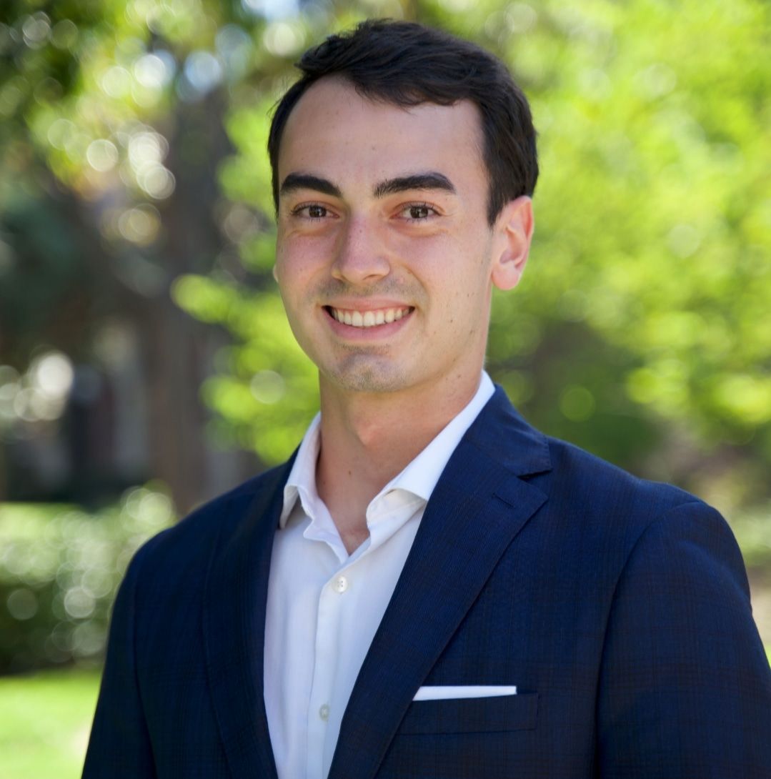 Man in navy suit and white shirt smiles outdoors.
