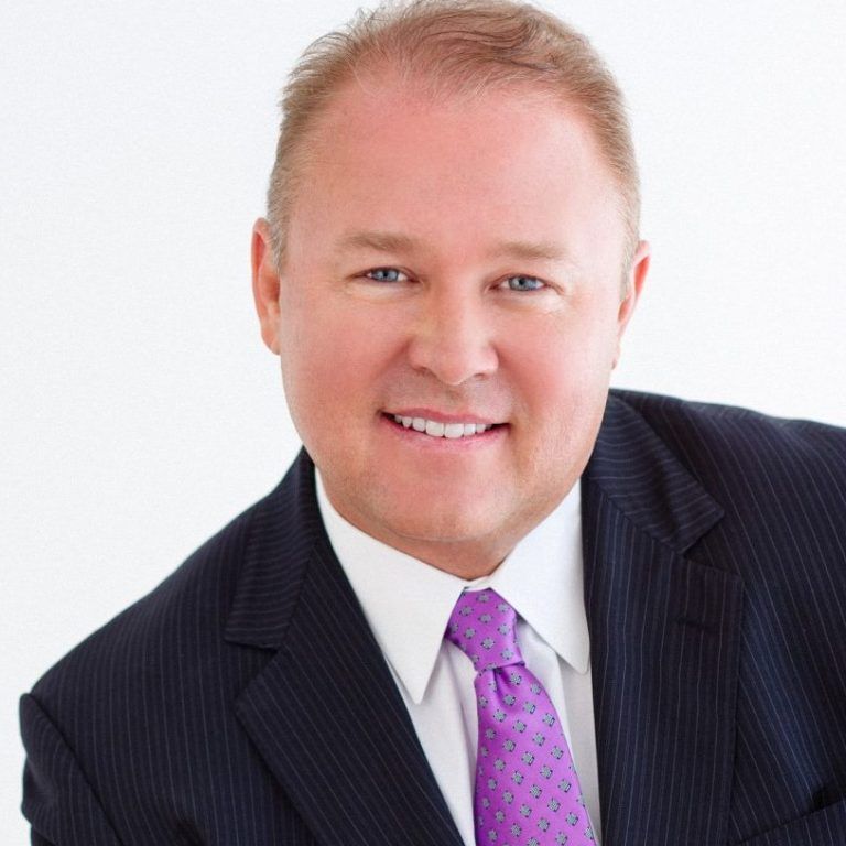 Man in a suit smiles, purple tie, against a white background.