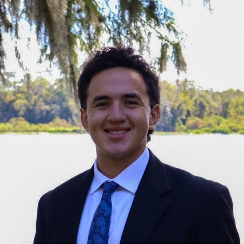 Smiling person in a dark suit and blue tie standing outdoors by a lake