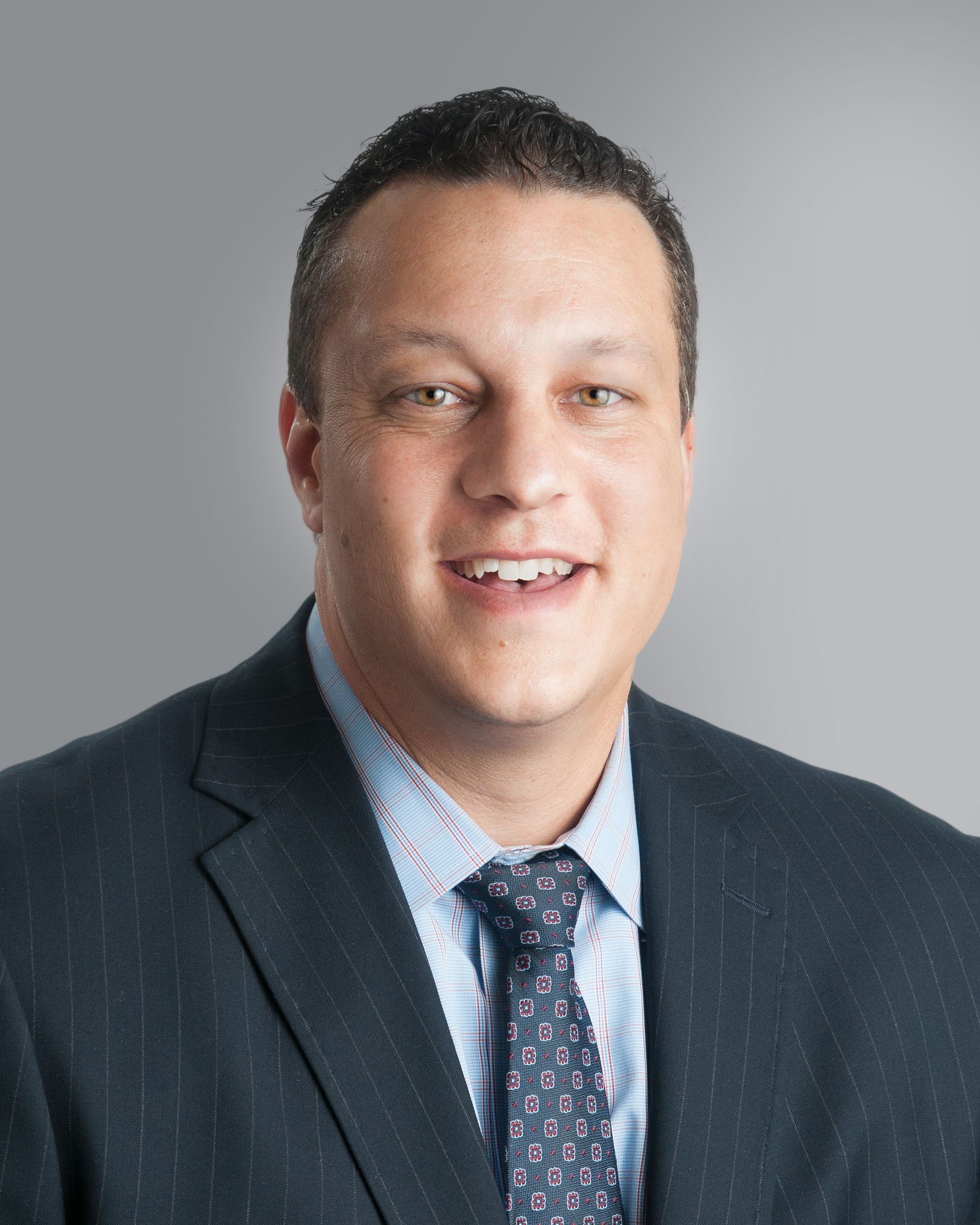 Man in a suit smiles at the camera, light blue shirt, patterned tie, against a gray background.