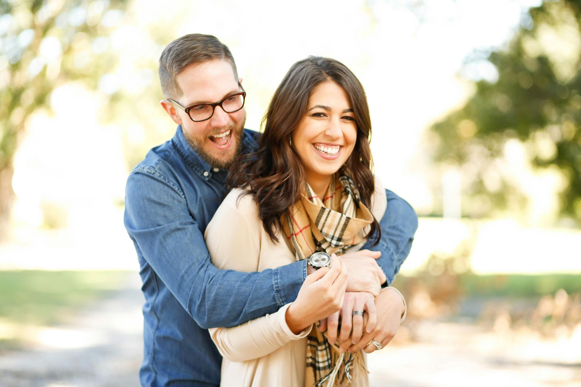 Man hugging woman from behind, both smiling outdoors. Beige and blue clothing, trees in background.