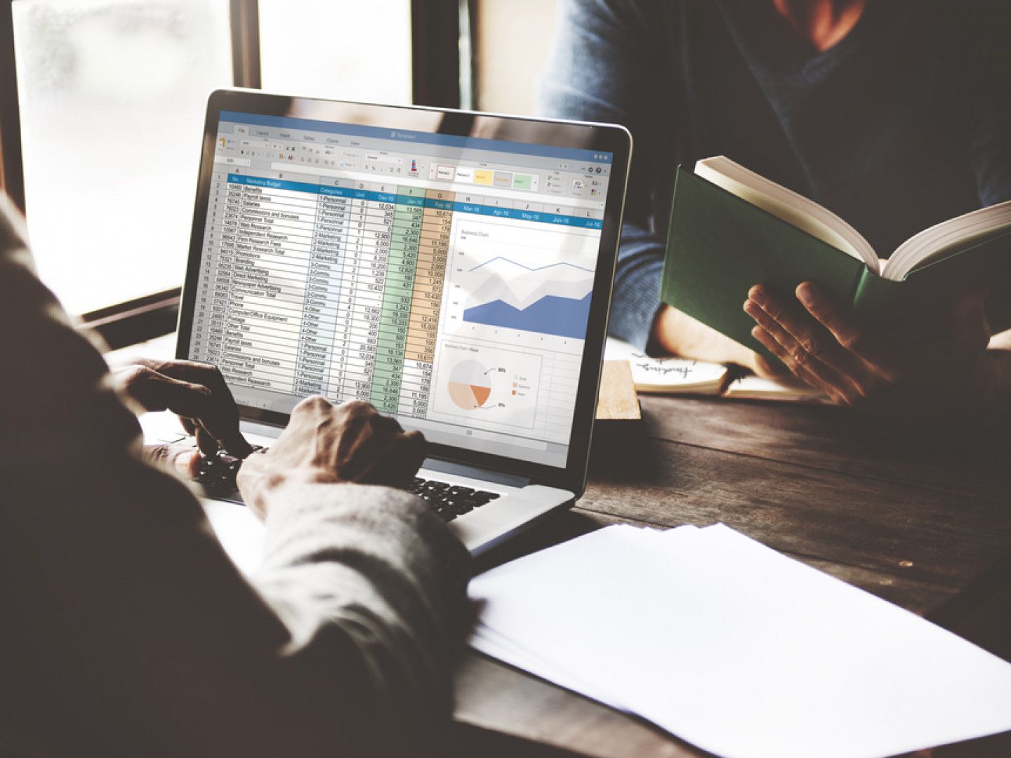 Person working on a laptop with spreadsheet data and graphs; another person reading a book.