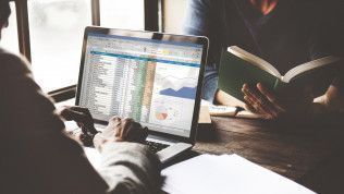Person using a laptop with a spreadsheet and charts, next to another person reading a book.