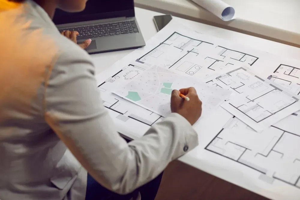 Person in a blazer, working on architectural blueprints at a desk with a laptop. — Adamson Town Planning in Noosaville, QLD