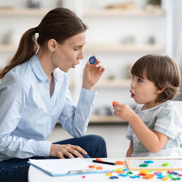 Child in a blue shirt talking indoors, facing a blurred person by a bright window.