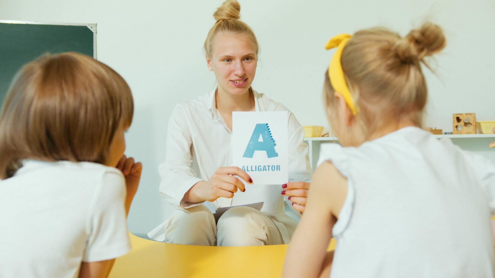 Teacher holding a blue letter A card while speaking to two children at a table in a classroom