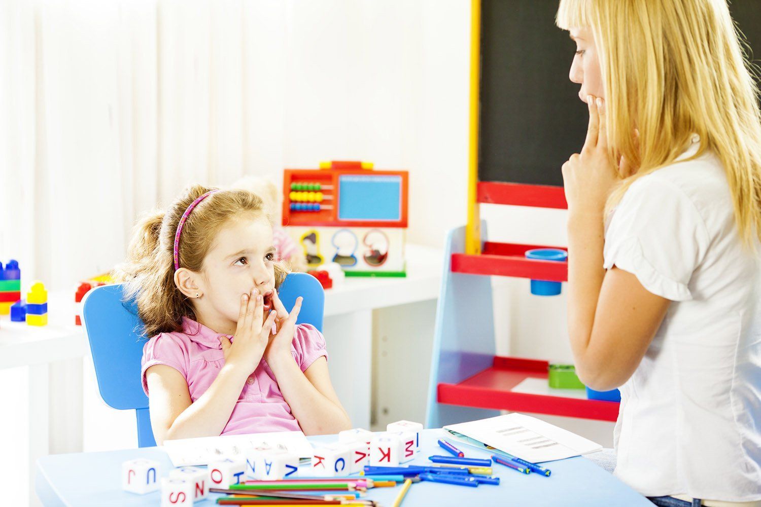 A woman is talking to a little girl who is covering her mouth with her hands.