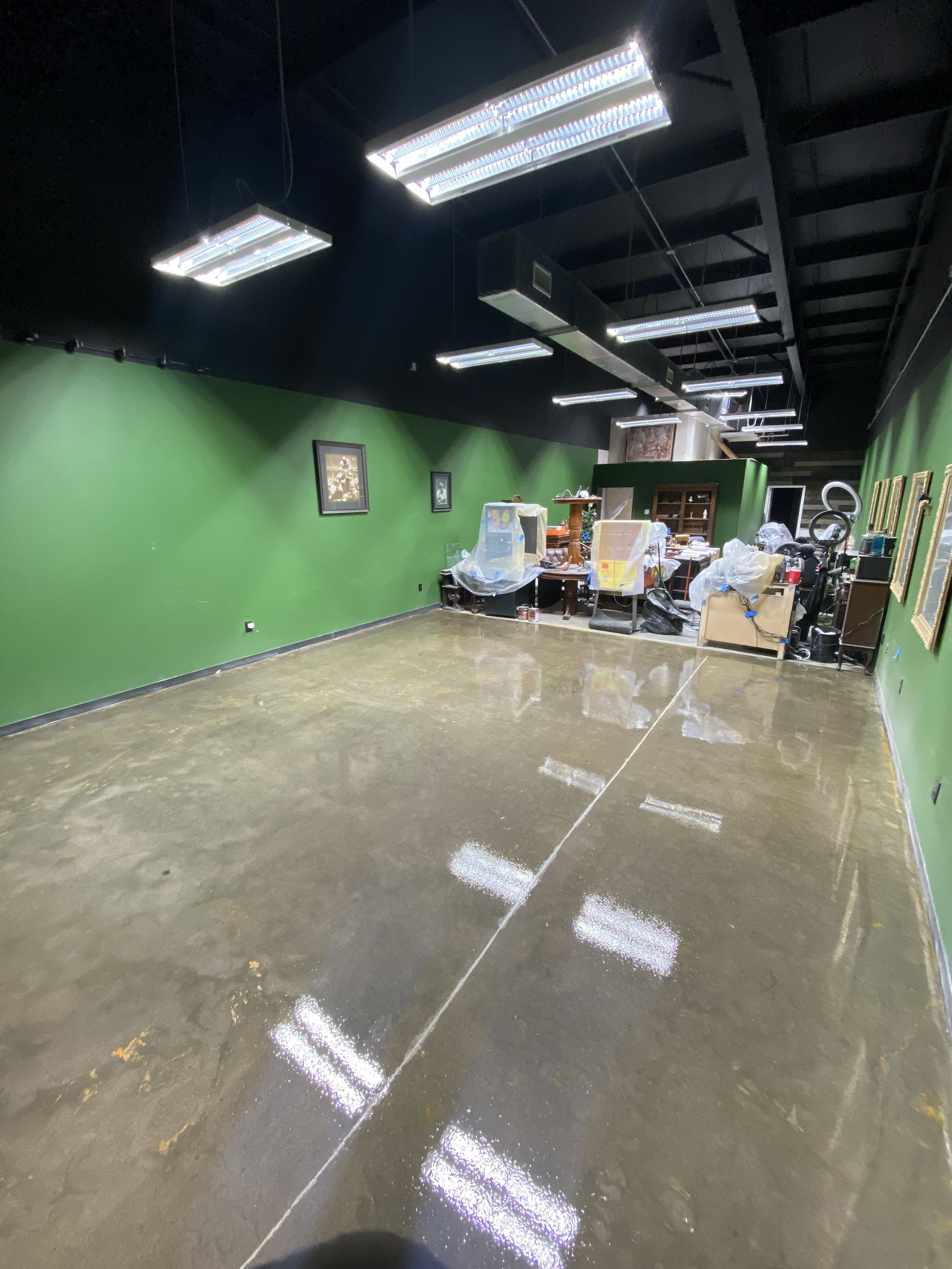 Empty room with green walls, black ceiling, and polished concrete floor. Lighting fixtures and clutter at the far end.