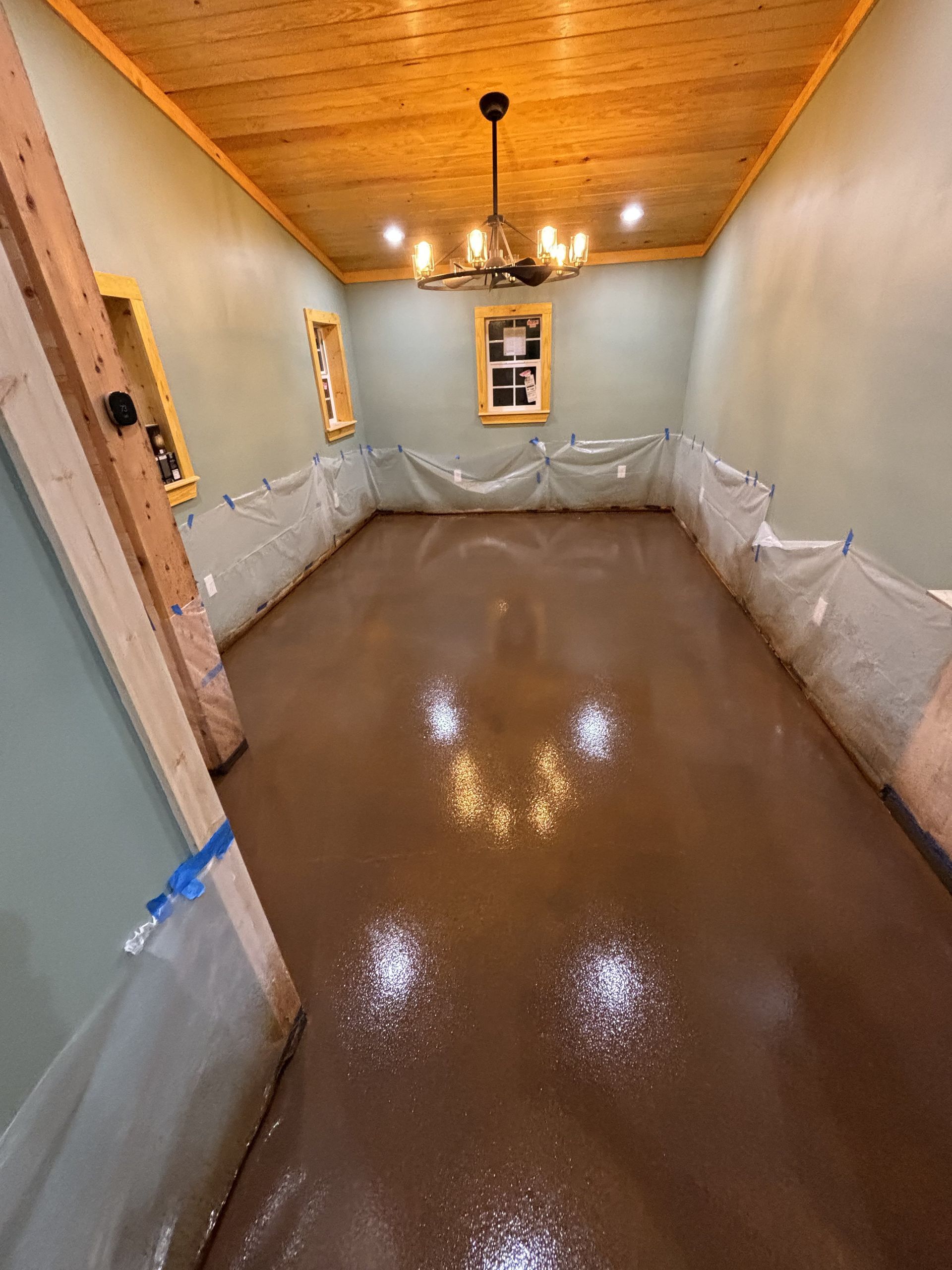 Room with brown floor, green walls, wood ceiling and trim, and chandelier. Protective plastic around perimeter.