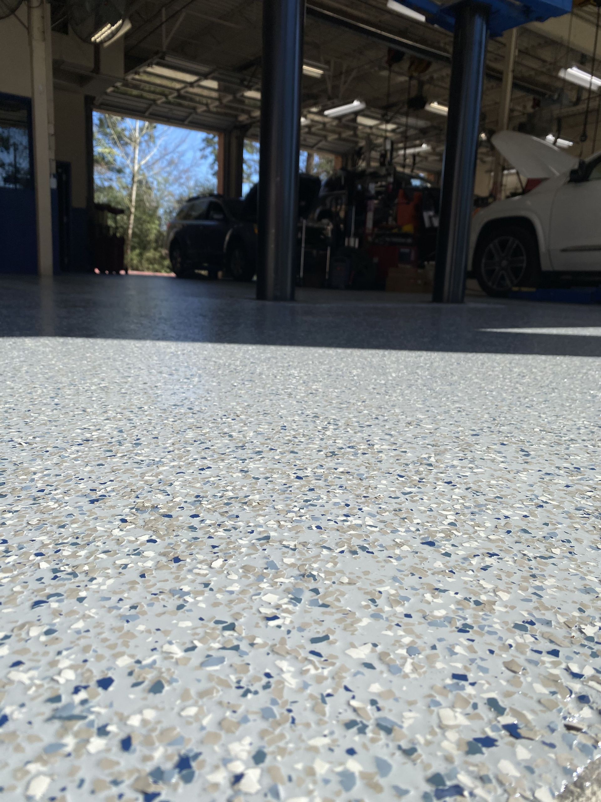 Garage floor with speckled blue, gray, and white epoxy. Cars and equipment visible in the background.