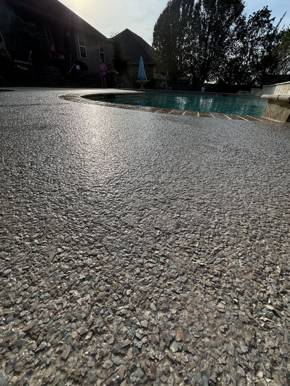 Gravel patio next to a pool with sunlight reflecting on the surface. Trees and a house are in the background.