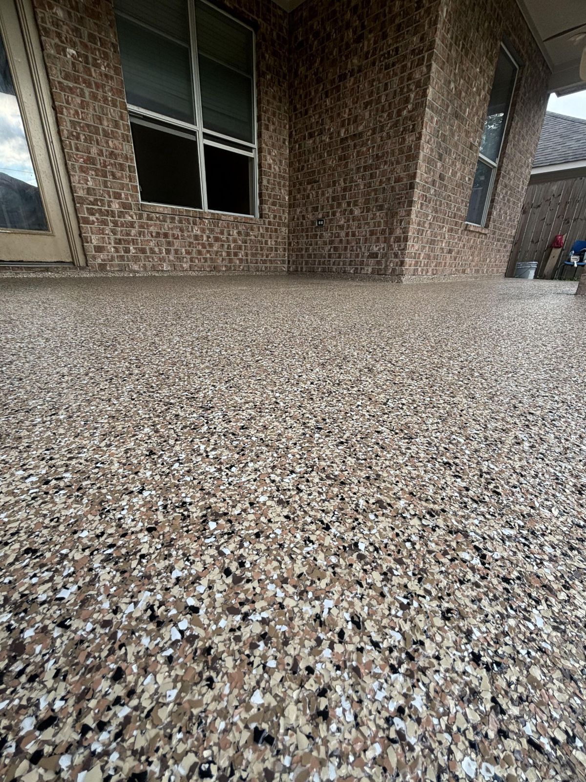 Epoxy-coated outdoor floor with beige, tan, and dark flecks, under a brick house with windows.