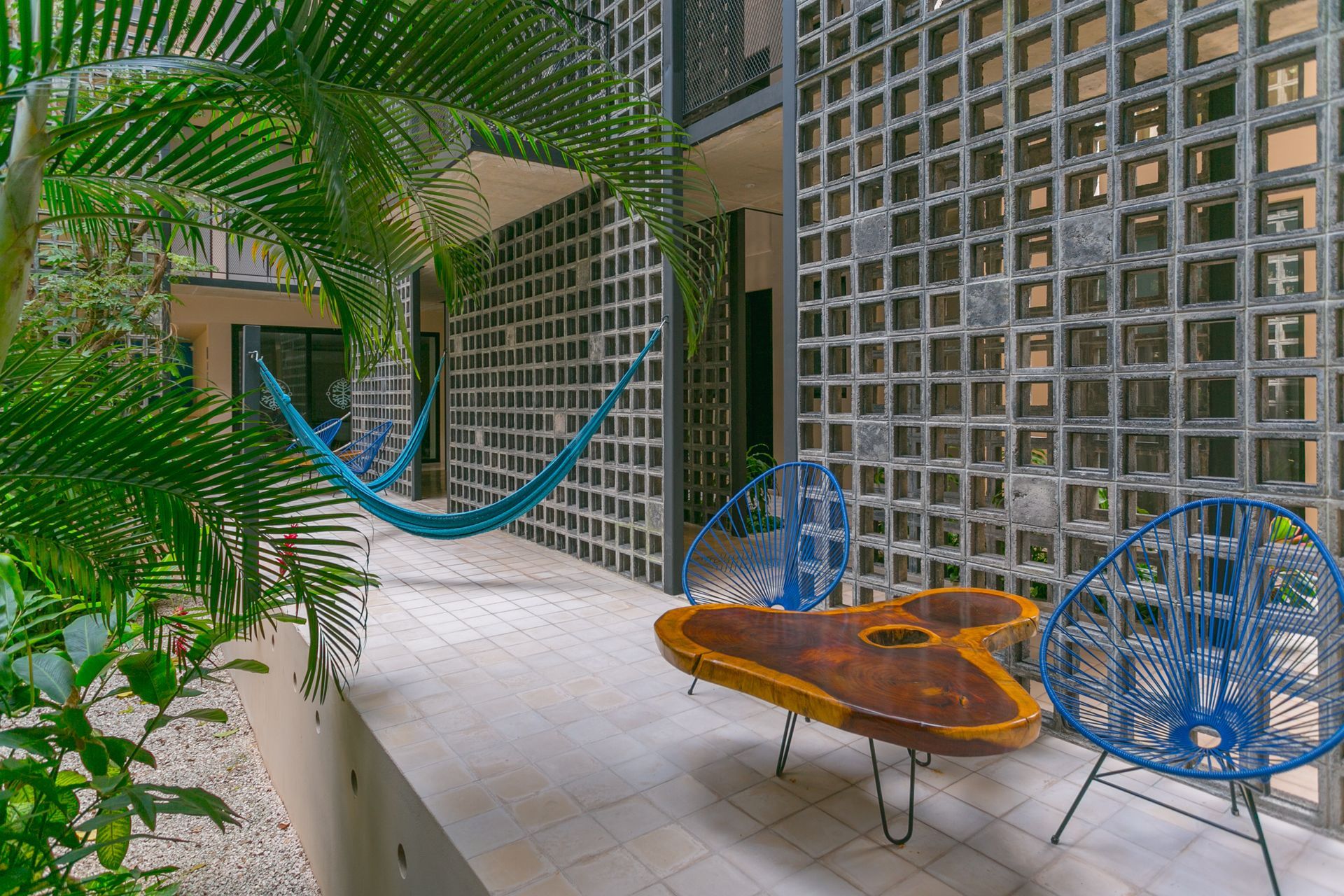 Patio con sillas colgantes azules, mesa naranja, suelo blanco y frondosas plantas verdes junto a paredes de bloques de vidrio
