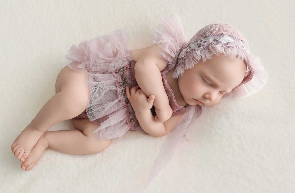 Sleeping baby doll in pink lace outfit and bonnet, curled on a cream blanket, holding a small teddy bear