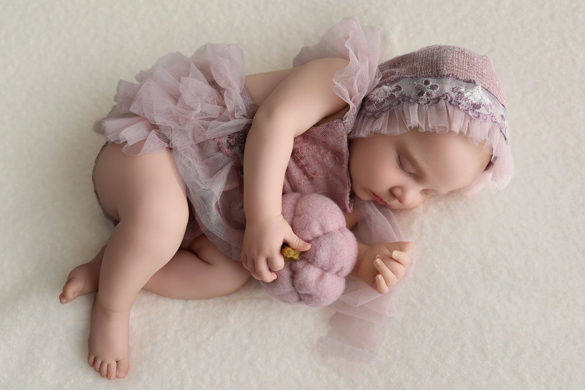Sleeping baby in a pink tulle outfit and hat, holding a stuffed pumpkin, on a light background.
