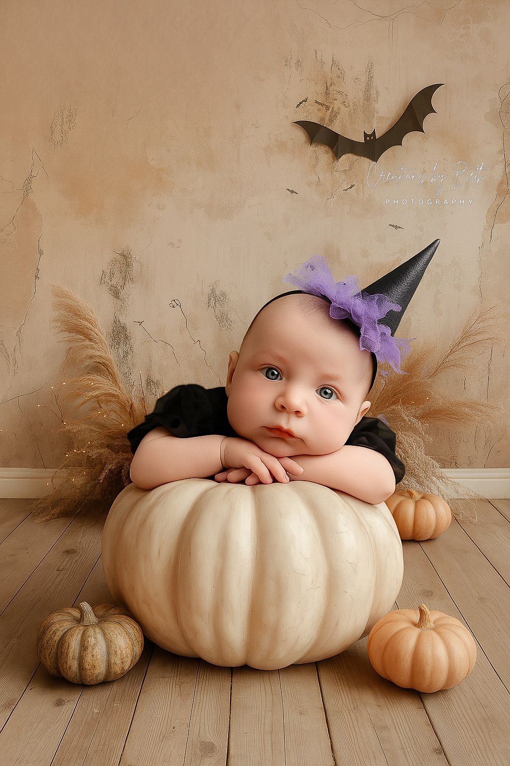 Baby in witch hat leans on white pumpkin, surrounded by pumpkins. Bat decor in background.