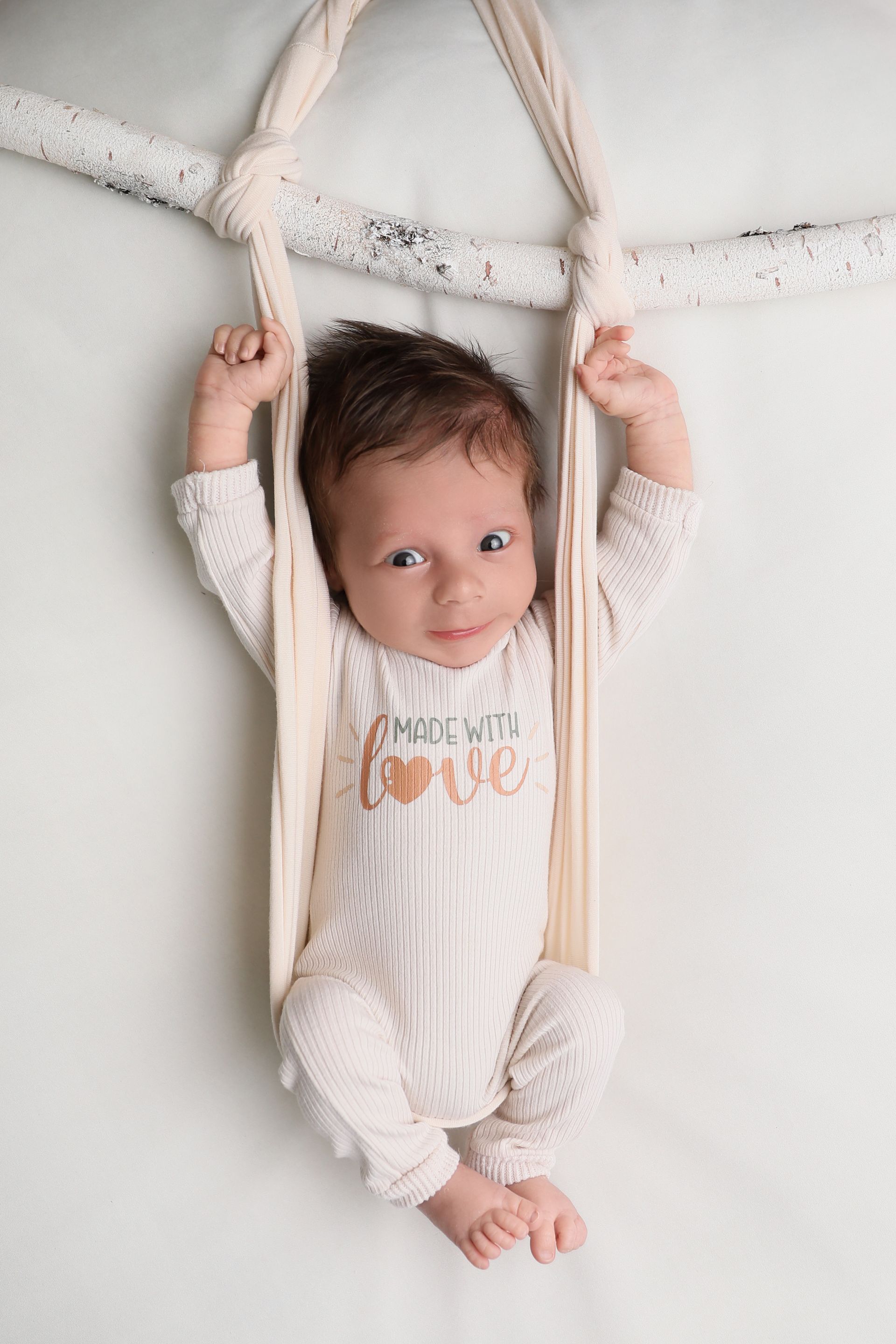 Baby in cream-colored onesie sits in a knotted swing, smiling. Background is light and neutral.