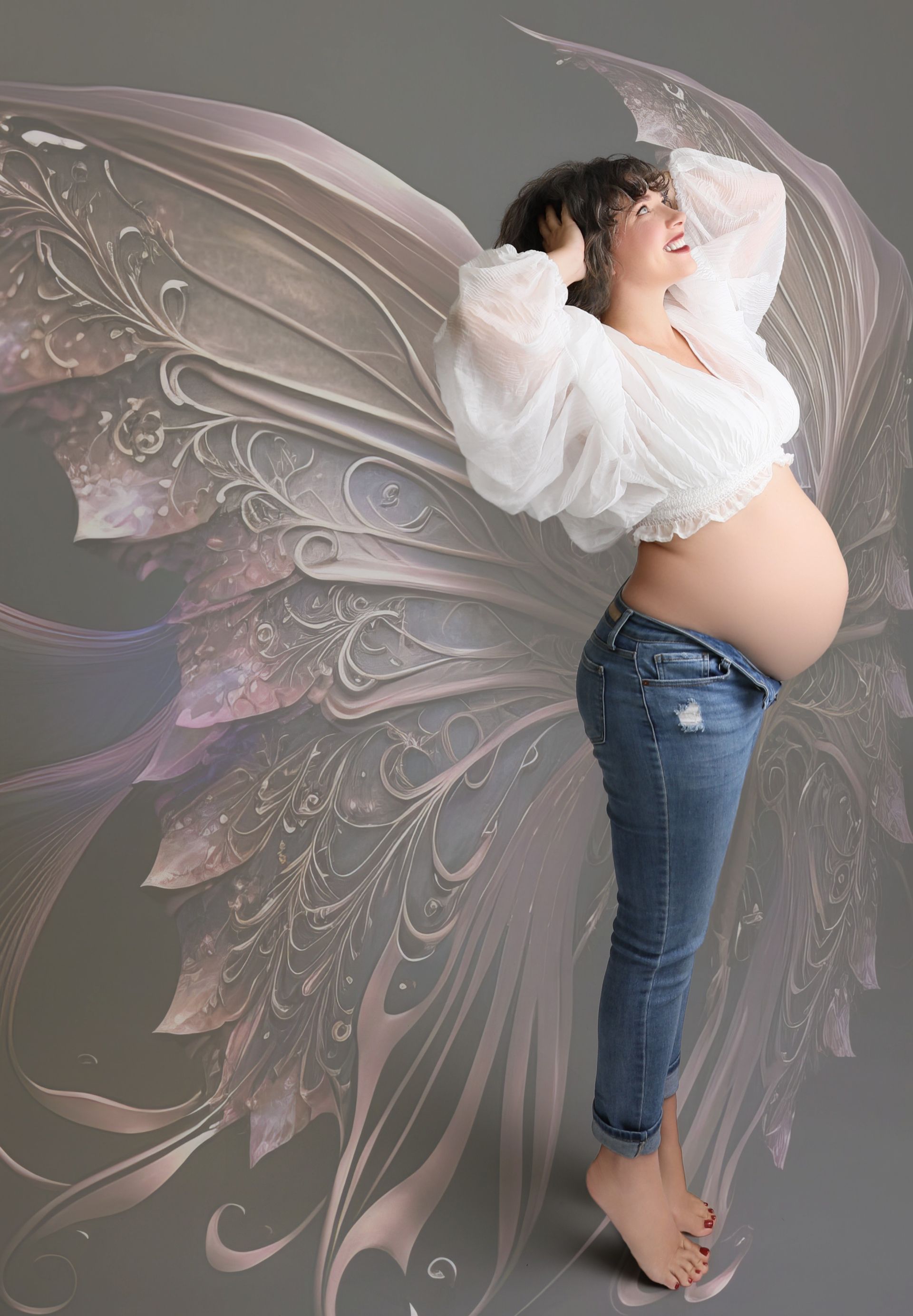 Pregnant person with fairy wings, arms raised, wearing a white top and jeans.