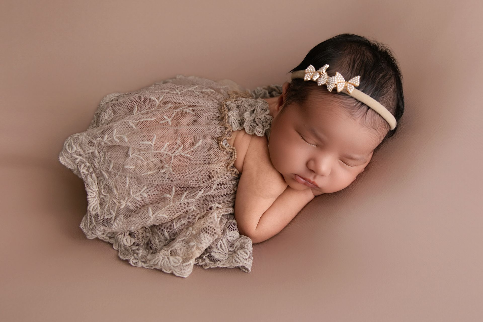 Sleeping newborn wrapped in lace, wearing a floral headband, resting on a beige backdrop.