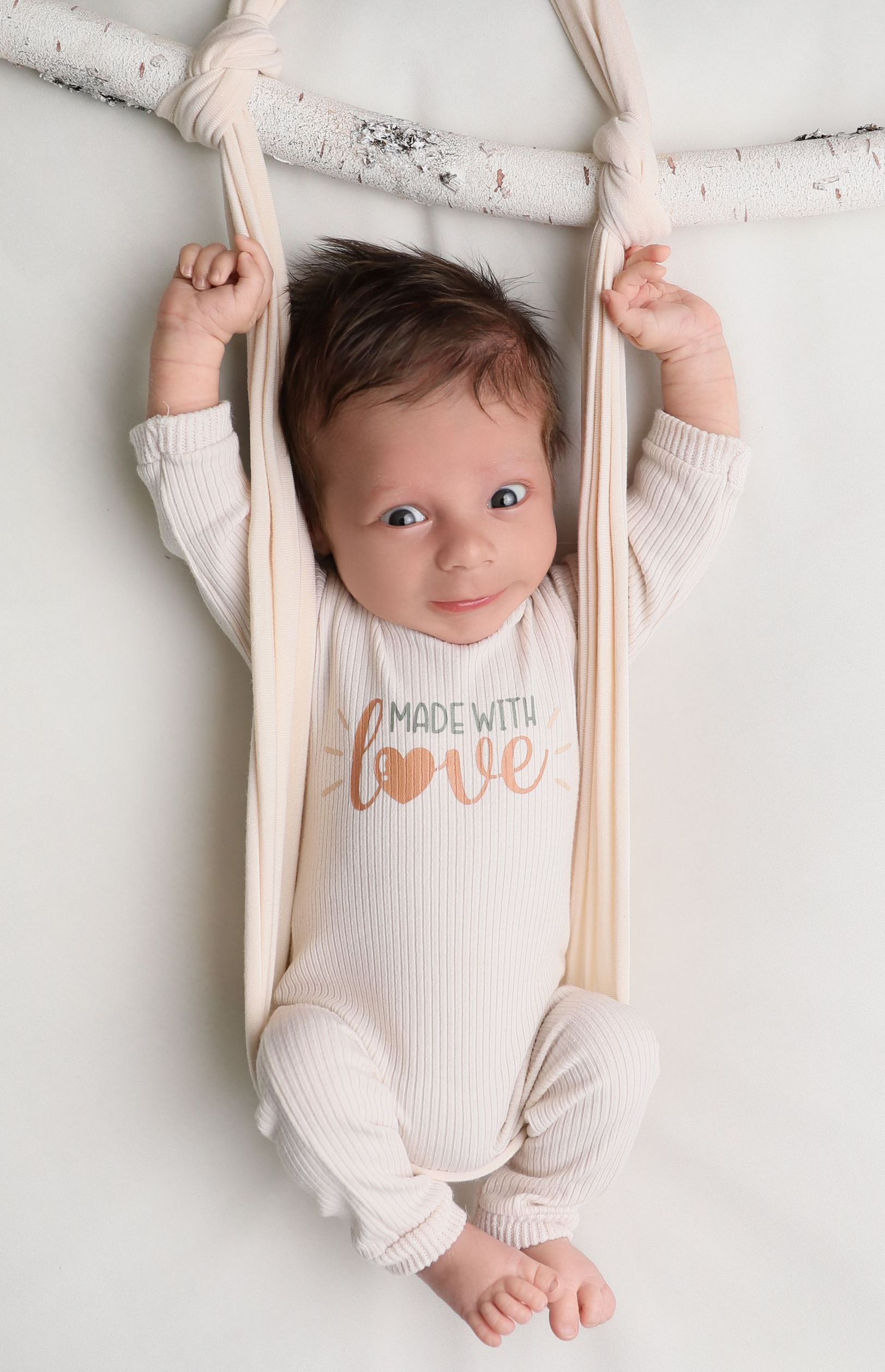 An awake newborn lies comfortably on a soft, white posing surface, gently supported with neutral fabric straps for safety. 