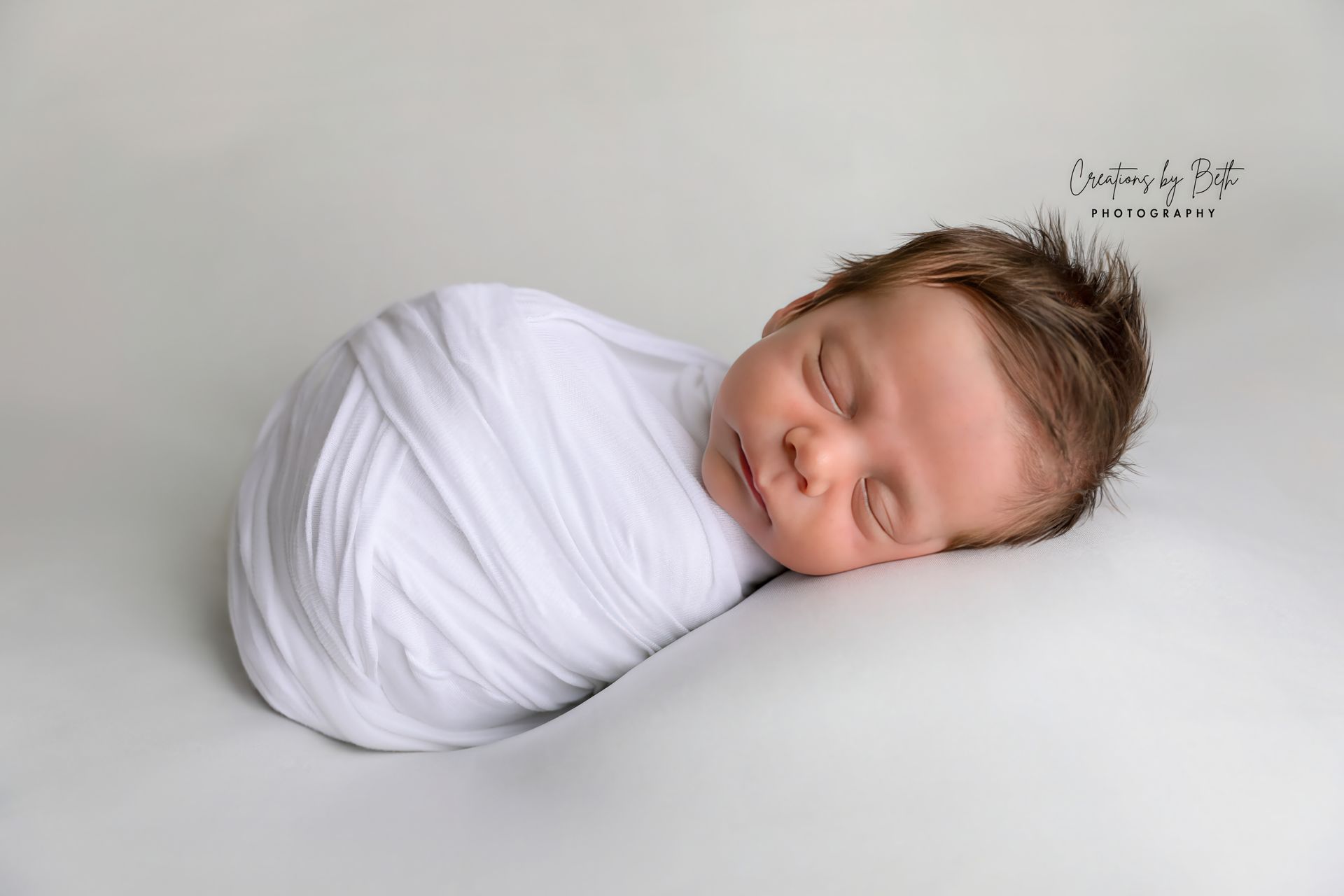 Newborn baby swaddled in white, sleeping on a white surface with light brown hair.