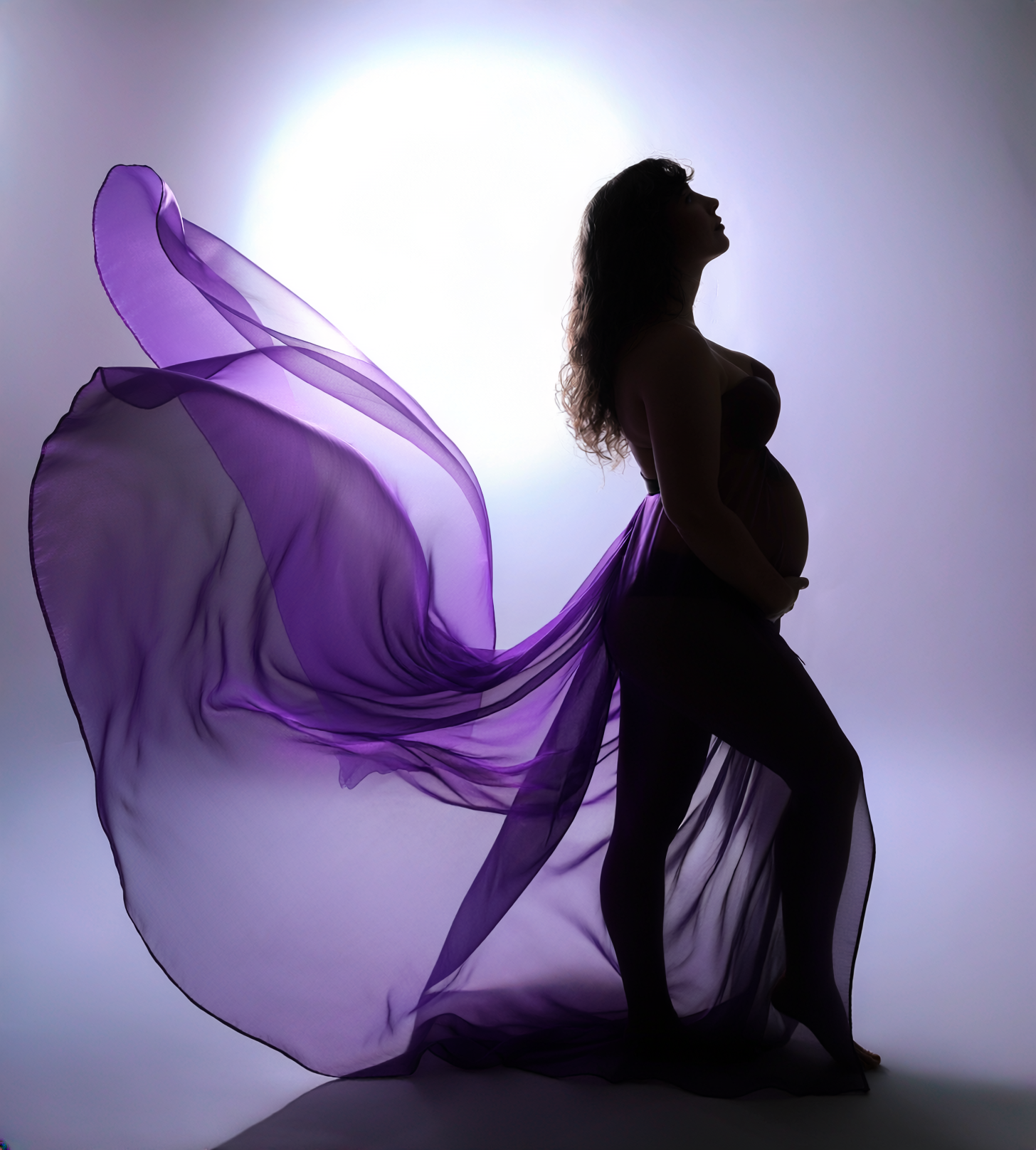 Silhouette of pregnant person, purple fabric flowing, hand on belly, looking upward.