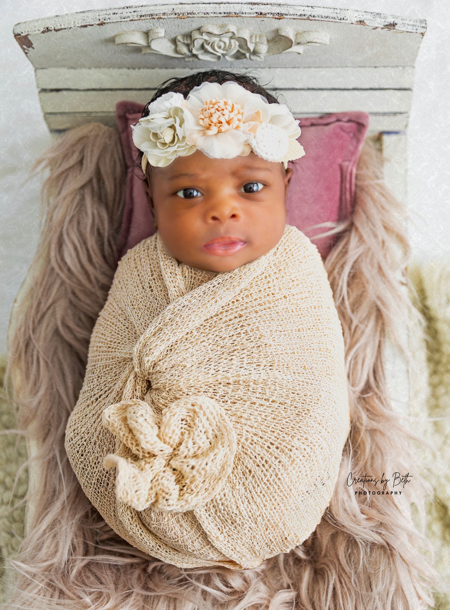Newborn baby swaddled in beige wrap, wearing floral headband, resting on furry surface.