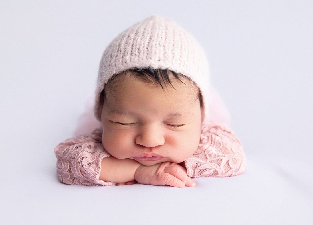 Newborn baby wearing a pink knitted hat and lace shirt, sleeping on a white surface.