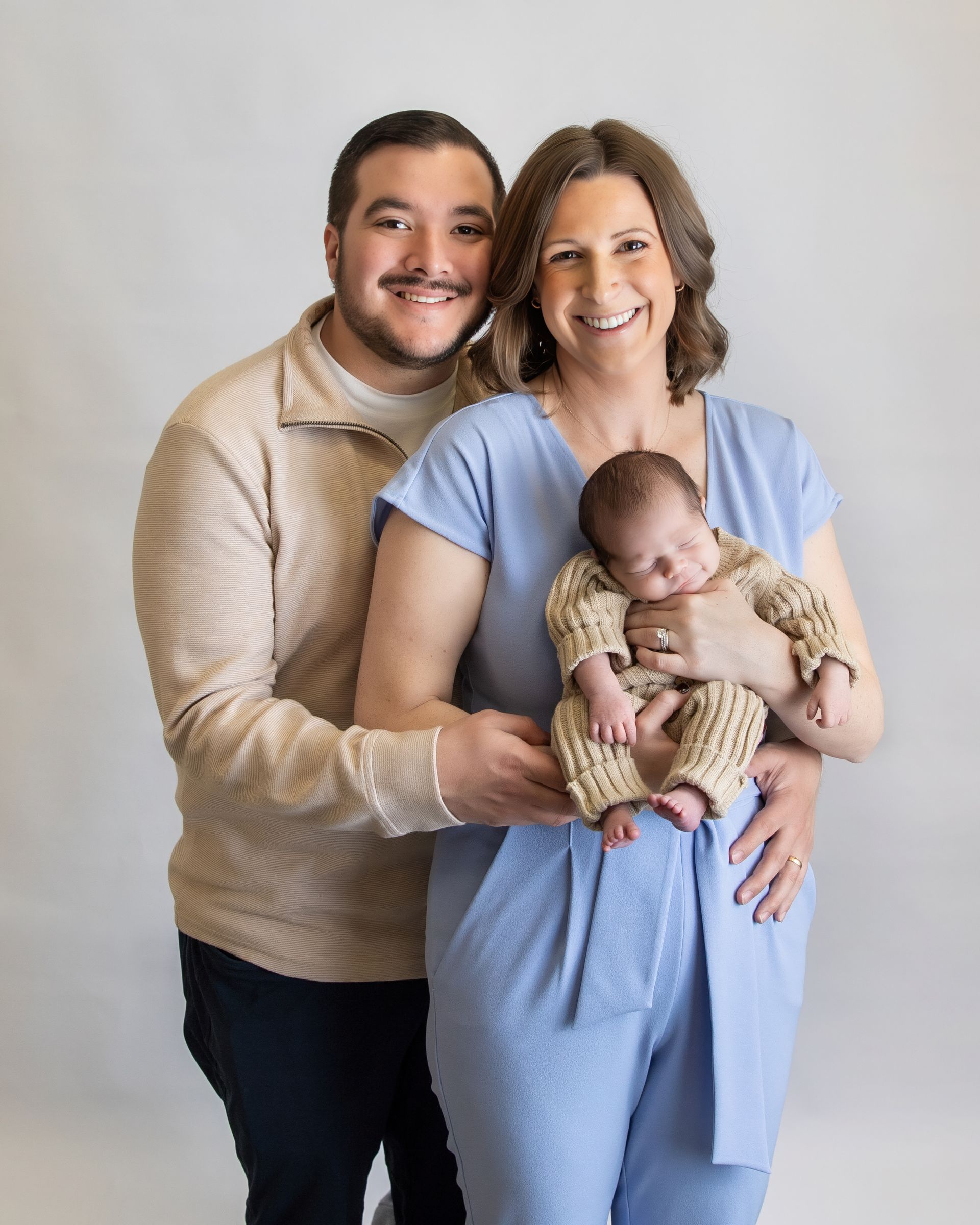 Smiling couple holding a sleeping baby, posed against a light gray studio backdrop.