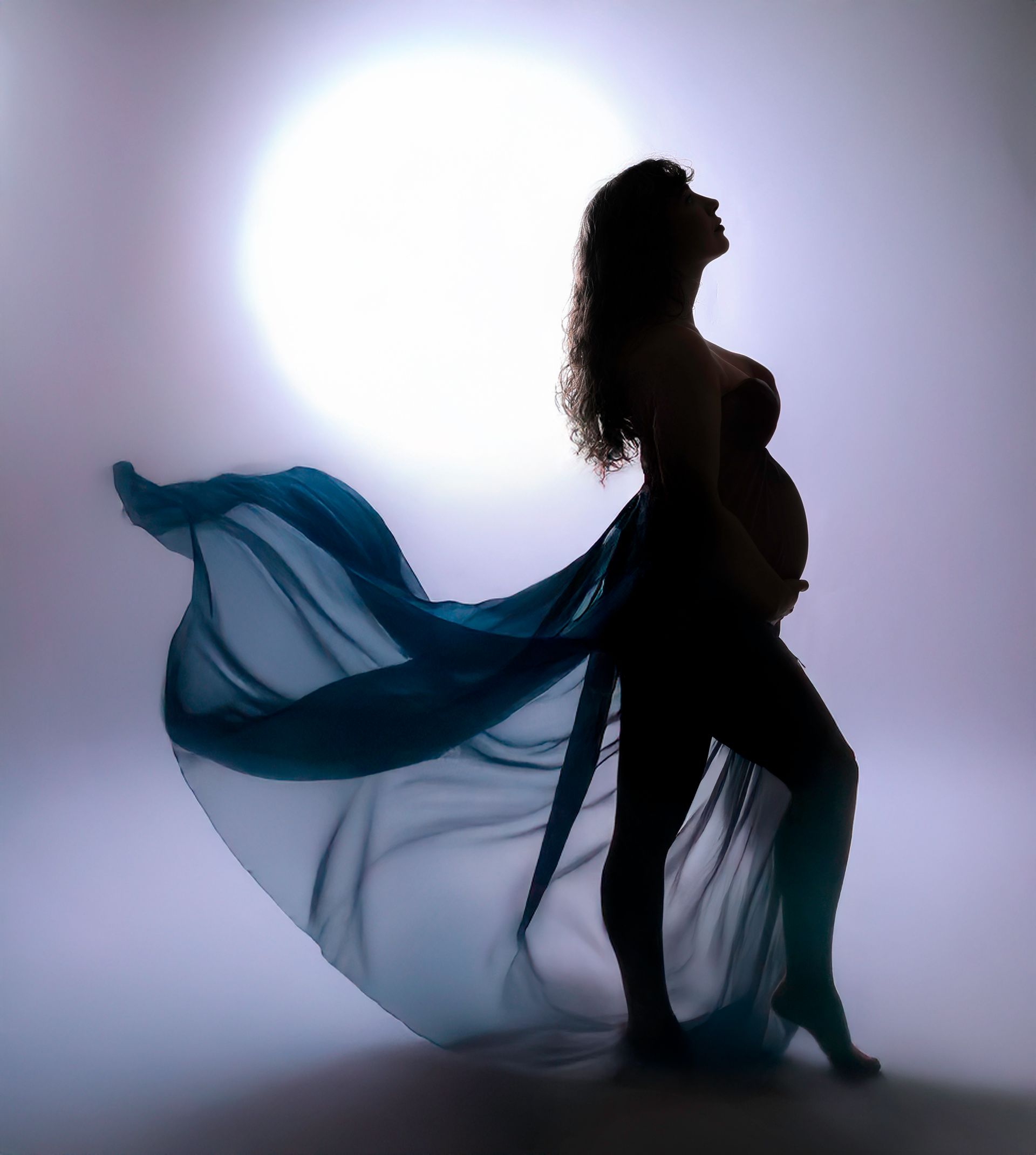 Silhouette of pregnant person in blue fabric, looking upward with hand on belly, against a bright background.