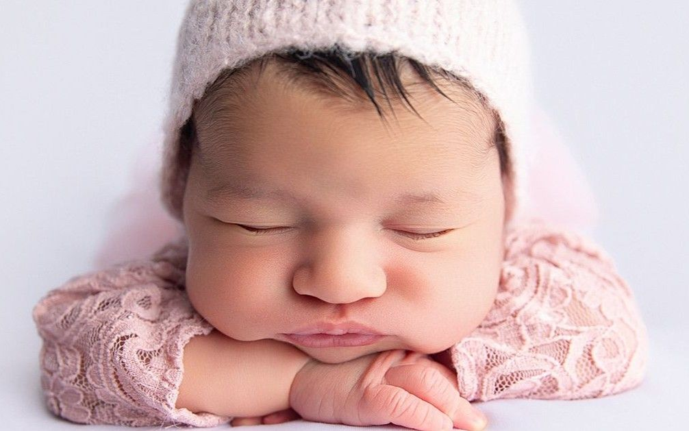 Newborn baby wearing a pink knitted cap and lace shirt, resting on hands, eyes closed.
