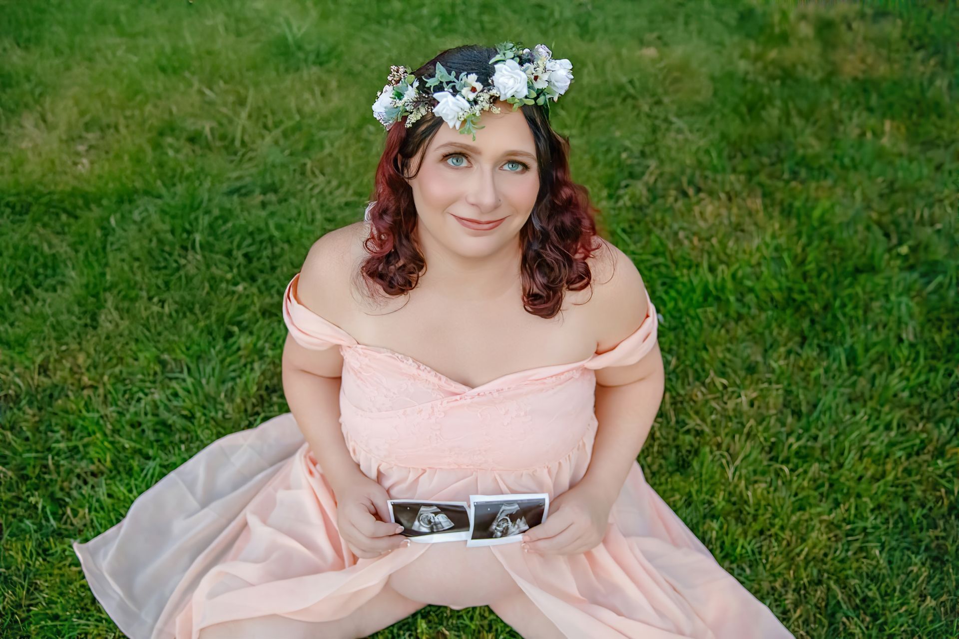 Pregnant woman in pink dress and flower crown, holding ultrasound photos, sitting on grass.