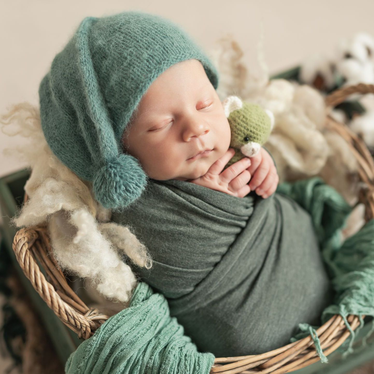 Baby swaddled in green, wearing a teal hat, sleeping in a basket with a small stuffed toy.