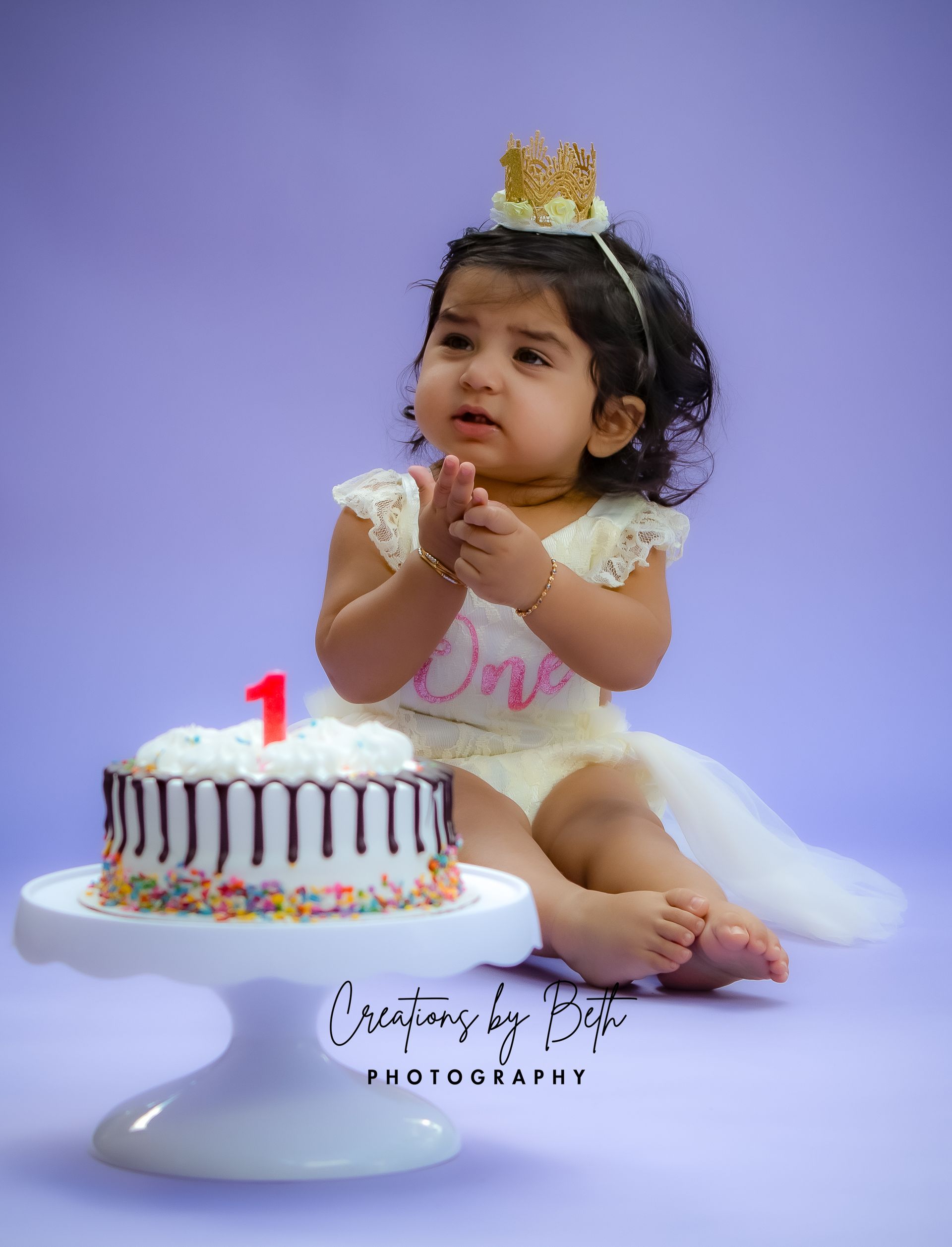 A baby sits next to a cake. Baby wears a crown and 