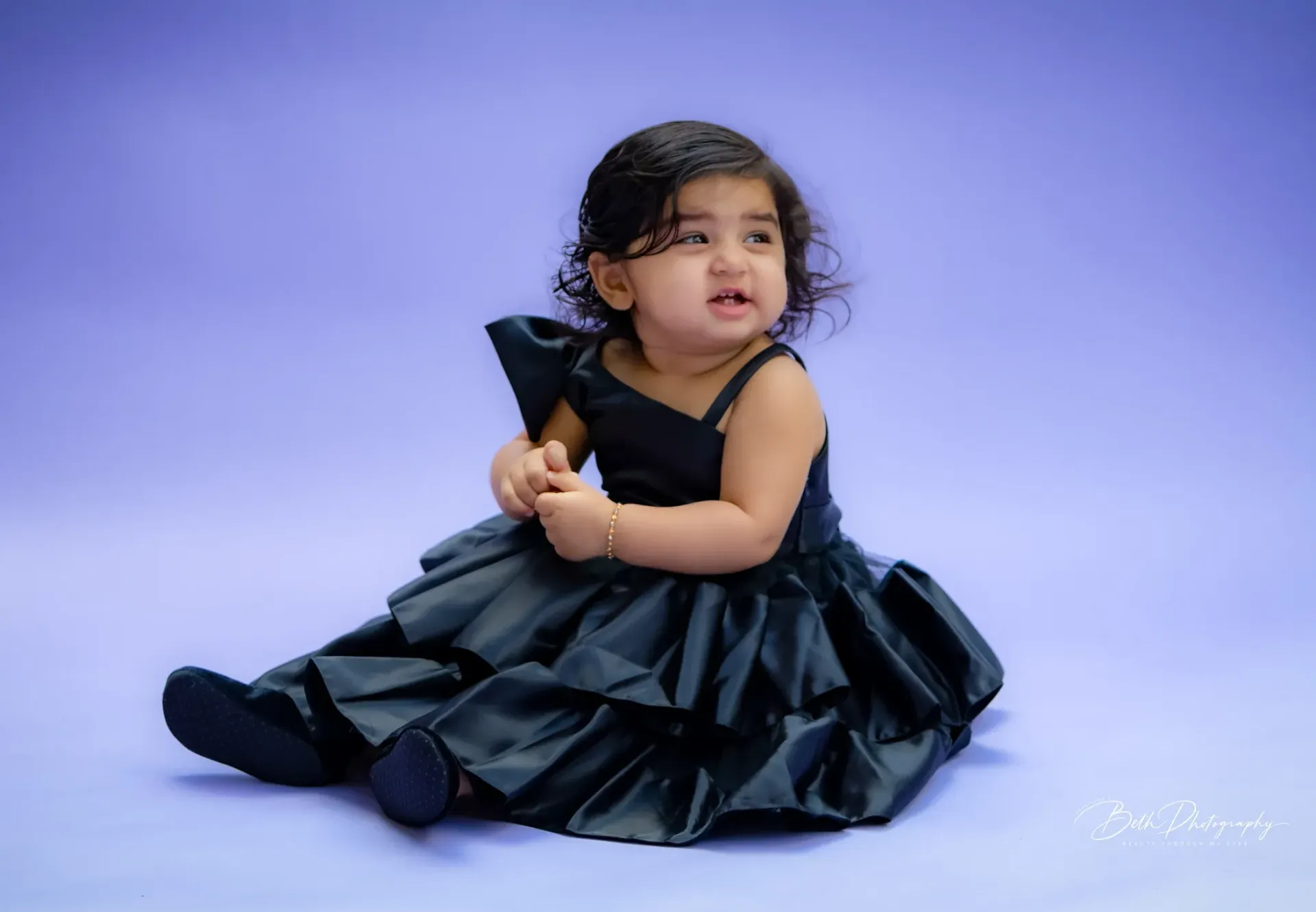 Child wearing a black dress sits against a purple backdrop, looking to the side.