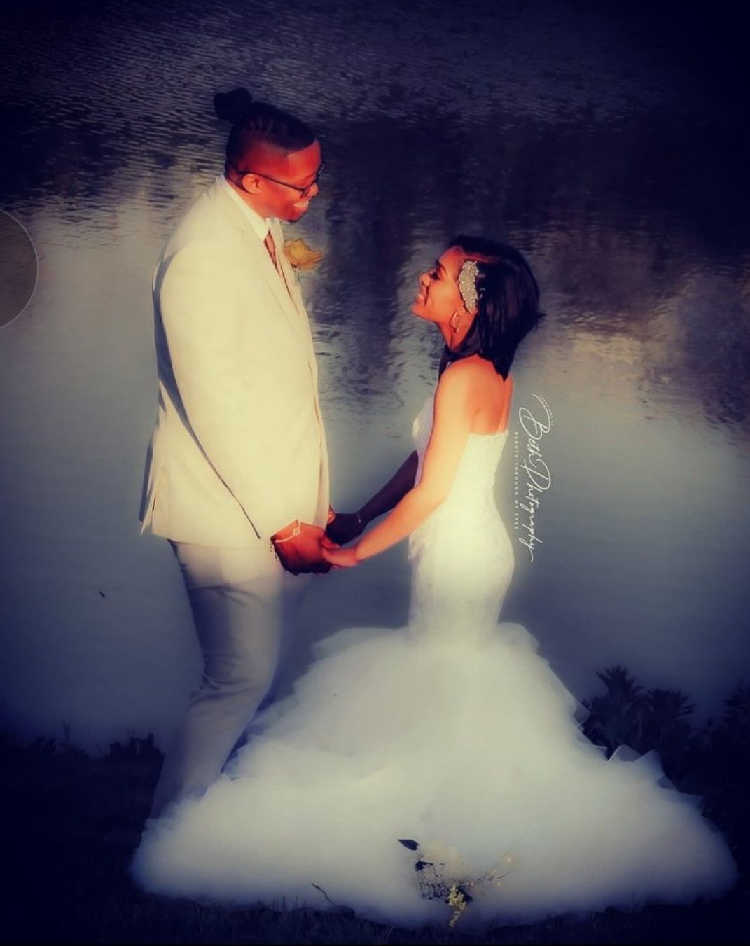 Couple in formal attire holding hands, looking at each other, in front of a body of water.