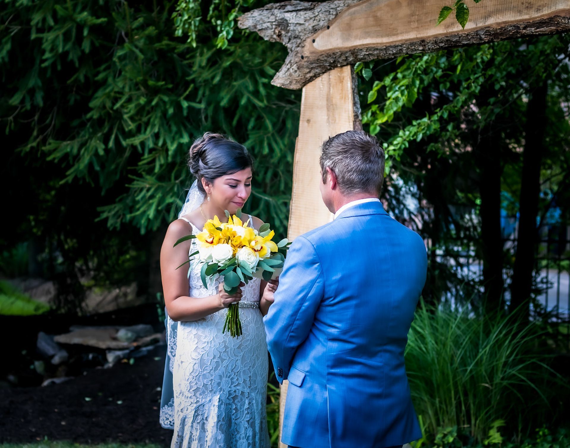 Bride and groom exchanging vows under wooden archway, holding flowers, outdoor setting.
